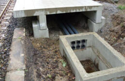 Vorgefertigte Stahlbeton-Aufbausch&auml;chte zur F&uuml;hrung von Strom- und Datenkabeln neben Bahngleisen, teilweise erdverlegt mit sichtbaren Kabelrohren.